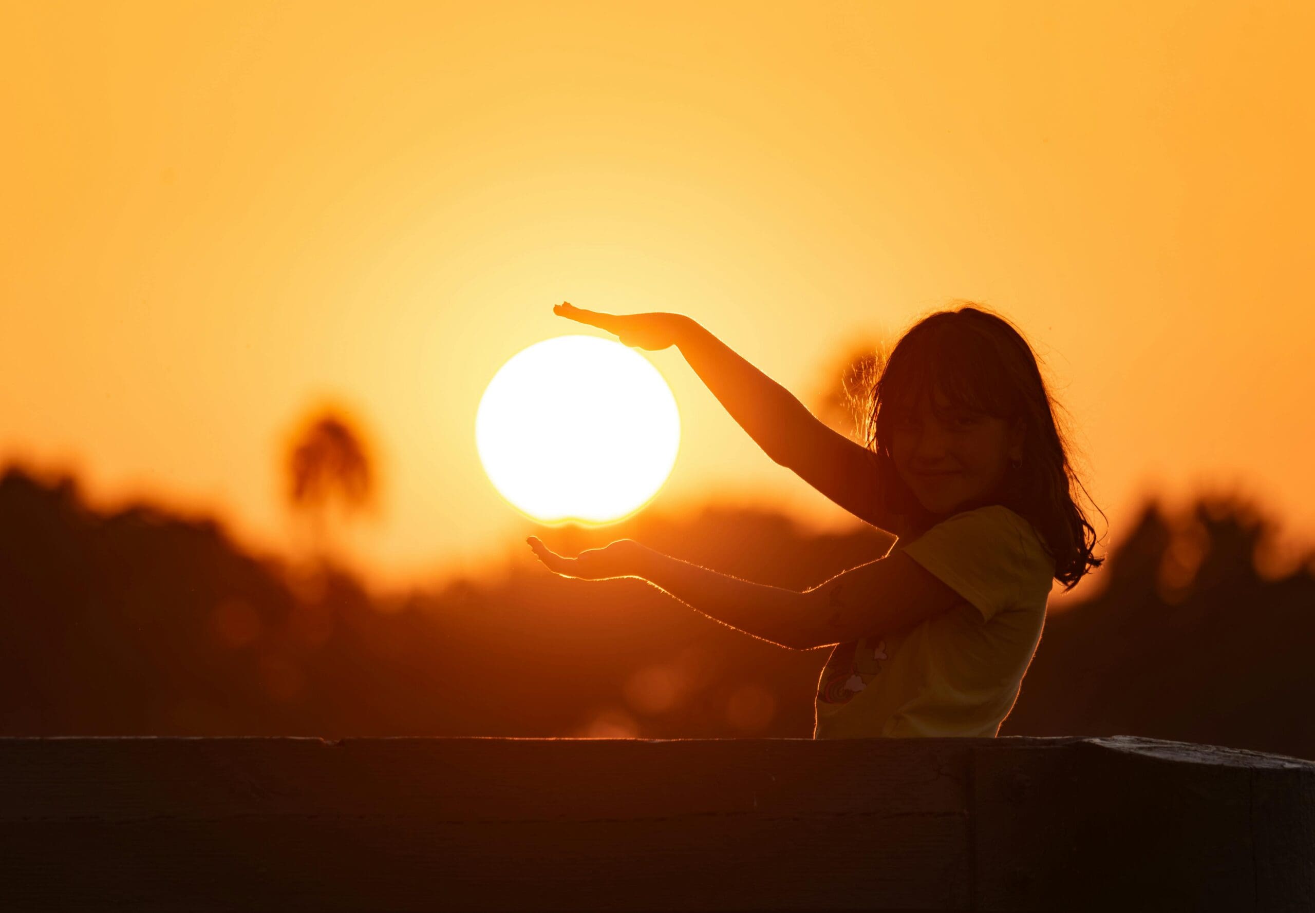 Smiling child playfully holding the sun at sunset, symbolizing joy, vitality, and vitamin D from sunlight vs supplements.